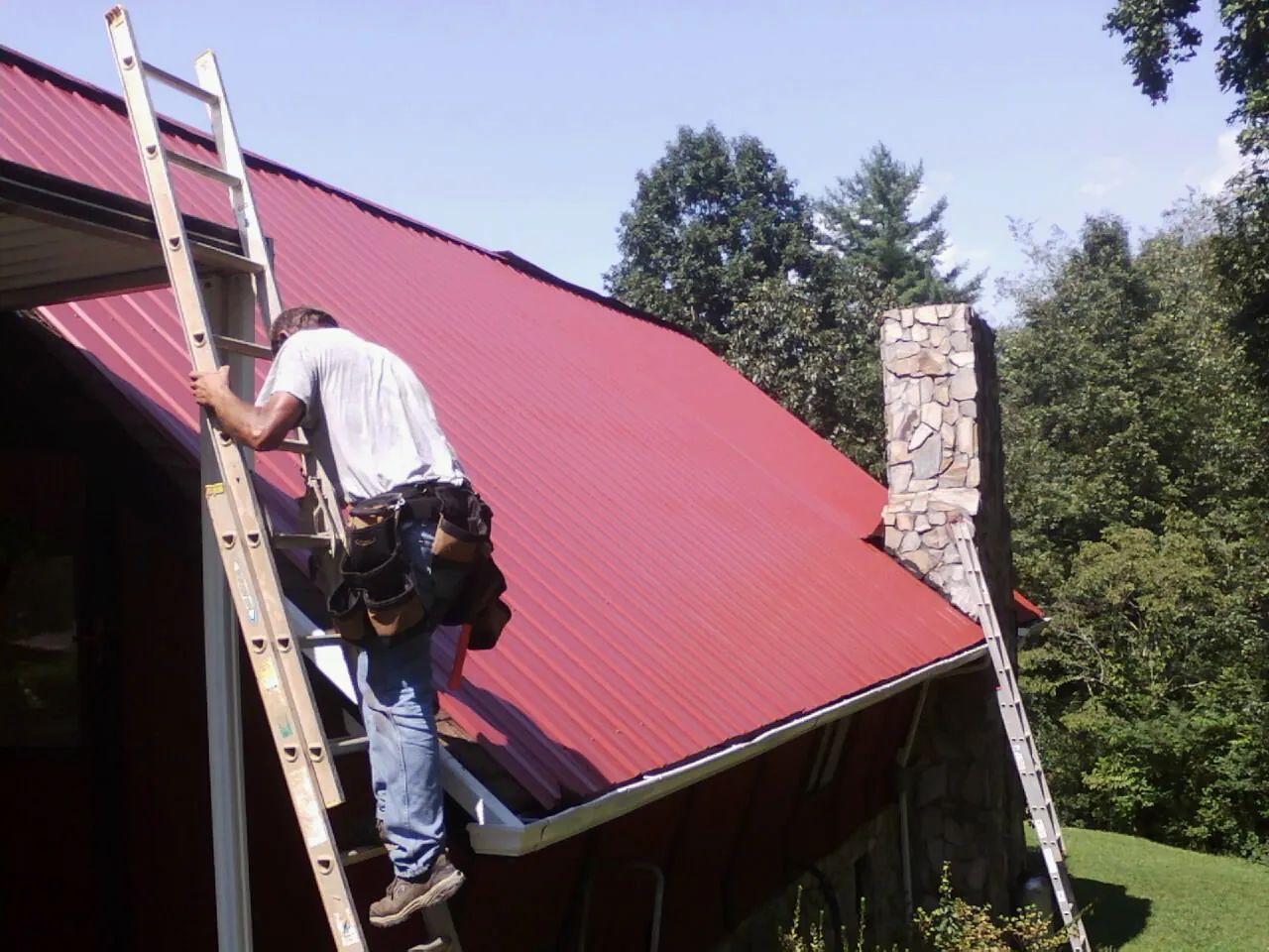 Roofer on a ladder beside a red metal roof and stone chimney — Soffit & Fascia Repair by Roofing Brothers in Wilsonville